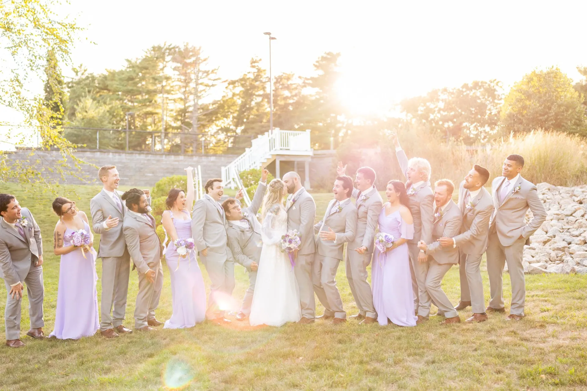 A bride and groom kiss at sunset surrounded by their cheering wedding party on the lawn at Rosebrook Event Center